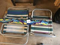 Two low beach chairs stacked, showing fabric reclining chair with blue and white stripes and mesh chair with multicolored stripes. Colorful umbrella wrapped and resting on chairs.