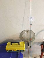 Fishing rod standing upright next to landing net and closed yellow plastic tackle box.