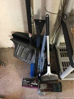 Two snow shovels leaning against wall along with three auto snow/ice removal tools, a manual battery charger in box, and a hydraulic jack in packaging on floor.