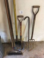Photo showing the full set of garden tools including shovels, pitch fork, hoe, and metal rake against a white wall on a concrete floor