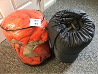 Two rolled polyfilled sleeping bags, one orange and one black, tightly packed and tied with straps