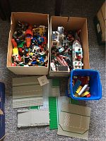 Two boxes filled with assorted vintage Lego and Duplo bricks, including vehicles and building pieces, along with multiple plastic Lego base plates showing road and grass designs.