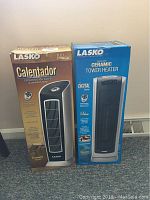 Two boxed Lasko ceramic tower heaters side by side, one in brown box and the other in blue box, showing front design and remote control functionality.
