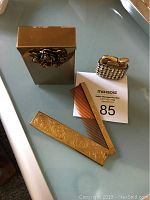 Front view of gold-tone cigarette case, foldable comb, and rhinestone decorated lighter on glass table