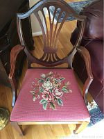 Front view of an antique wood dining chair with a needlepoint floral seat cover in reddish-pink fabric, featuring carved wood back and armrests.