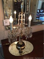 Pair of antique brass candelabra electric lights with crystal prisms on etched glass round mirror trays placed on a wooden surface, bulbs lit.