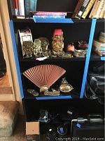 Brass decor items on black bookshelf including bookends, napkin rings, paperweights, fan-shaped decor, and small bowl; displayed with some books around and blue painter's tape on shelf edges.