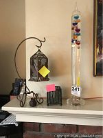 Image showing the 24 inch Galileo thermometer and two metal candle holders on a white mantel against a wall with art partly visible.