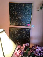 Two square botanical art pieces with gold frames shown hanging on a pink wall, photographed with some pink flowers and part of a lamp visible.