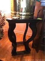 Black wooden side table with decorative curved legs and rectangular top, placed on hardwood floor with some faux floral arrangements nearby
