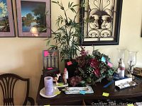 Wide view of the lot showing faux flowers, candle, figurine, wooden box and other decorative items on a table with framed wall art in background.