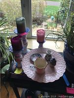 Overall display of pink cake plate, three candle holders with candles, Lenox vase, and small ceramics on table by window with plants.