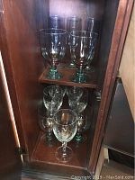 Four larger green-tinted stemware glasses on upper shelf with five smaller matching glasses below on lower shelf in wooden cabinet.