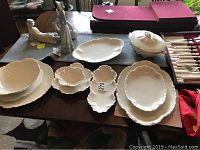 White porcelain serving pieces arranged on table, scalloped edges, different sizes and shapes, one covered dish with handle.