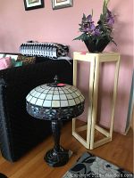 Full view of stained glass lamp and lacquered plant stand with mirror top beside black couch and pink wall