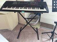 Image showing Casio CTK 550 keyboard on black X-style stand with carpeted floor background. Partial view of black metal music stand to the right.