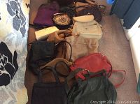Photo showing overall assortment of 9 ladies purses in various colors and styles placed on carpeted floor near bed.