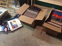 Two cardboard boxes and loose stack containing vinyl LP records on concrete floor