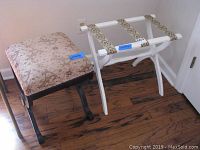 Wood bathroom bench with plush fabric seat and curved legs with gold accent medallions next to white folding luggage rack with tapestry bands.