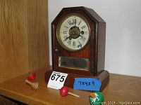 Front view of William Gibbons wooden mantel clock with visible glass door and round face with Roman numerals, sitting next to small colorful wooden spinning tops on a wooden shelf.