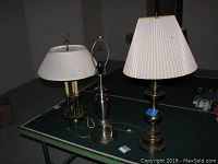Three table lamps displayed on a table, including a brushed stainless steel lamp and two brass lamps with pleated white lampshades.