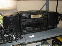 JVC black boombox radio with two detachable speakers and cable coiled beside it on shelf.