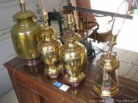 Four brass table lamps on a wooden table, showing different shapes and sizes. All have wooden bases and brass bodies with bulb holders and electrical cords.