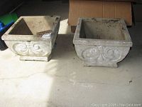 Two square cement planters with scroll embossed design, placed side by side