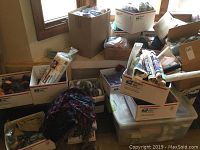 Wide view displaying multiple boxes, bags, bins containing yarn and fabric supplies, including USPS Priority Mail boxes and a large plastic bin with lid.
