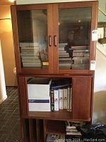 Wooden media storage cabinet with glass doors and books inside, showing vertical handles, and lower section with sliding wood doors and vinyl record slots.