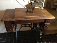 Wide view of vintage Singer sewing machine table with wood surface and black metal frame and pedal, with miniature sewing machine model on top.
