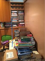 Wide view of stacked books and some packed in boxes, showing a large collection primarily of cookbooks and novels.
