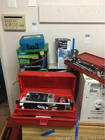 Red metal tool box open with assorted wrenches, sockets, and small hardware pieces inside. Additional items clustered behind including tool boxes and packaging.