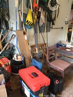 Wide-angle view of garage corner showing garden tools like rake, limb trimmer, red and black toolboxes, chair, and various hanging cords.