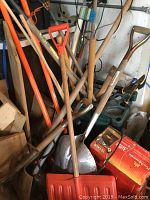 Photo showing multiple garden tools including rakes, shovels, and the orange red plastic snow shovel, also visible wooden handles and metal blades/tines.