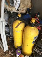 Photo showing two yellow scuba tanks with black valve handles and visible scratches and soiling on tanks surface.