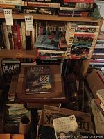 Bookshelves filled with assorted books including crafting guides and fiction novels, stacked with additional books on floor and table.