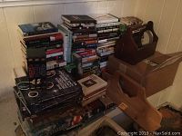 Photo showing multiple stacks of books on the floor featuring various titles, wood box with handle, and the wooden shelf