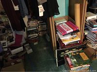 Photo showing stacks of various books including craft and knitting books, children's books, and posters scattered around.
