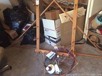 Full view of two wooden wool winder frames standing on floor, with a metal and plastic swift yarn winder loaded with yarn cones on carpet in front. Background contains boxes and other items in storage.