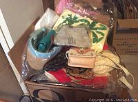 Photo showing assorted purses, makeup travel bags, evening bags, and totes piled on top of a cardboard box, some in plastic wrapping.