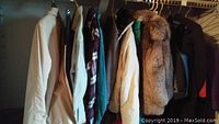 Closet view showing a variety of men's jackets and coats including beige trench coat, fur-lined plaid jackets, denim jacket, and other outerwear hung on a rod