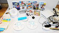 Photo showing Nintendo Wii console, multiple games, white steering wheels, Wii remote with blue cover, white speaker accessory, and various cables including sensor bar and AV cables.