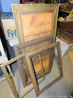 Multiple large wooden and gold-tone picture frames stacked against a wall in various sizes showing ornate designs, condition details with visible aging.