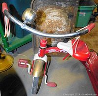 Overview of tricycle showing handlebar with bell and red tassel handles, part of seat and front wheel with metal fender.