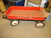 Side view of vintage red Rex wagon showing rectangular red metal body with branding and white wheels with rust spots, missing pull handle