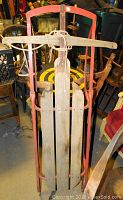 Front view of vintage sled with red metal frame, wooden slats seat and attached wooden steering bar with rope.