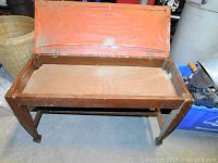 Photo showing the piano bench with the storage lid open, displaying the interior storage space and the worn orange cushioned seat lid.