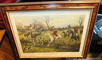 Framed hunt scene art showing riders on horses and hounds in a rural setting, framed in a wooden frame with glass.