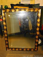 Front view of rectangular mirror showing black wood frame decorated with ornate gold leaf floral and vine pattern along all edges.